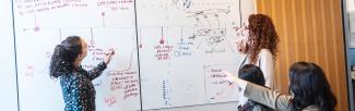 Two people write on a whiteboard while another points at something